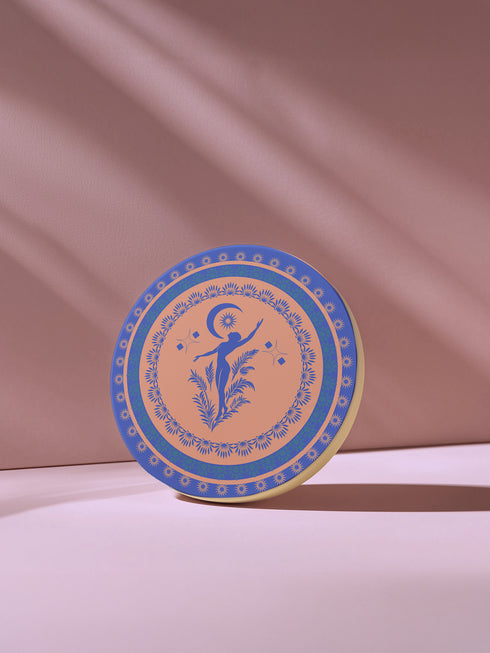 Round ceramic coaster featuring a silhouetted dancing figure reaching toward a crescent moon, surrounded by concentric rings of stars and floral patterns in blue on peach background